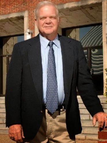 A man in a suit and tie standing in front of an entryway to a building made of brick and doors made of glass aided by meta...